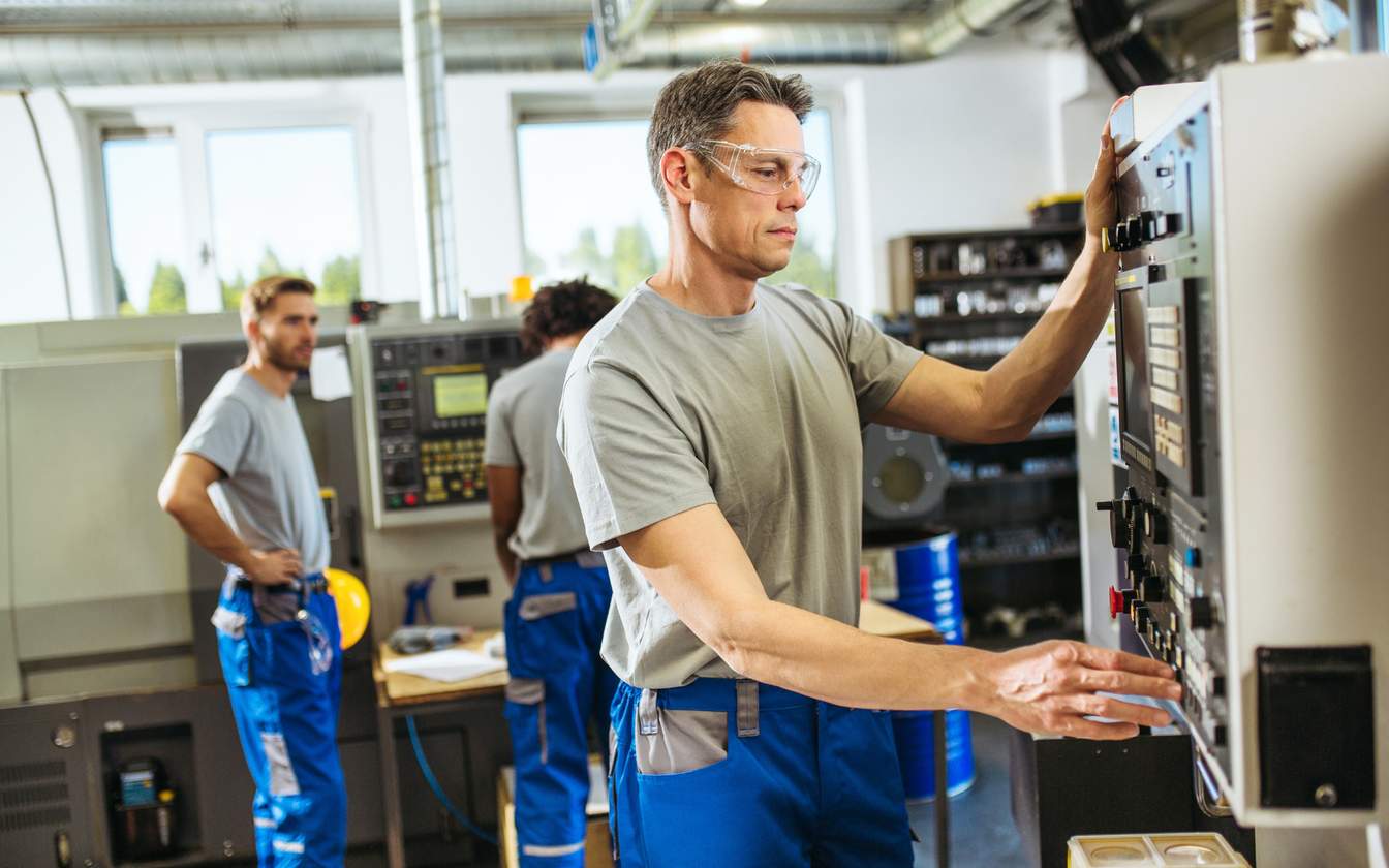 3 male engineers wearing grey shirts and blue work pants and safety glasses operating a machine